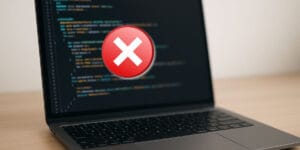 foto de um notebook com a tela desfocada com código de programação e um simbolo de erro no meio da tela