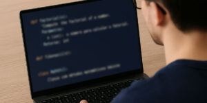 Pessoa programando em um notebook, vista de trás, com código desfocado exibido na tela em um fundo escuro