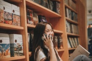 Side view of young cheerful ethnic female with book speaking on cellphone in public library while looking away