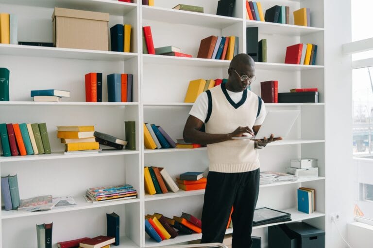 African American man using a laptop in a well-organized library setting with colorful bookshelves.