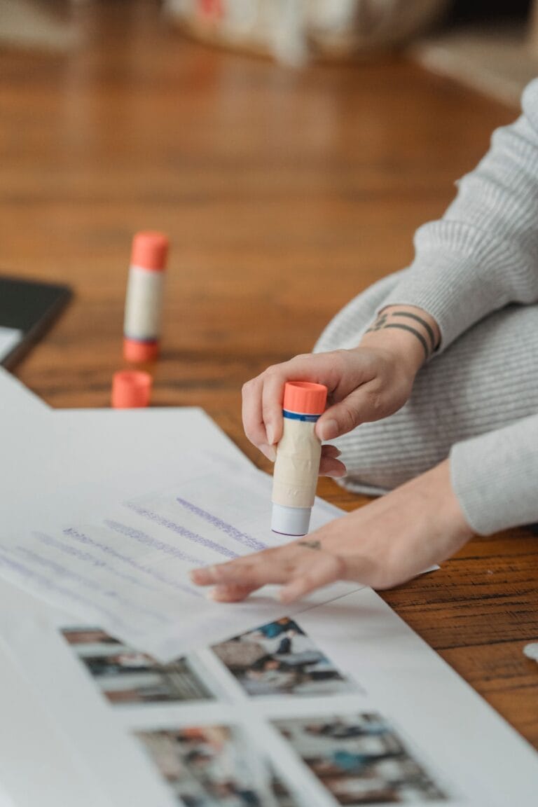 Crop unrecognizable person in gray sweater applying glue stick on paper while forming photo album on floor