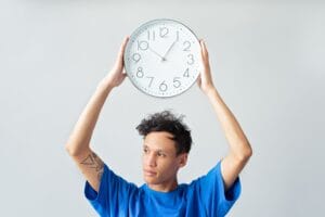 A man in a blue shirt holding a wall clock above his head, contemplating time.